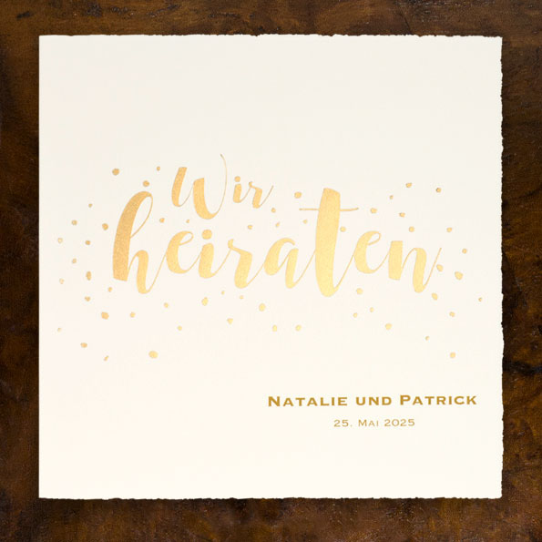 Vergrößerte Abbildung Bütten-Einladungskarten zur Hochzeit, mit goldener Schrift. Goldene Konfettis wirbeln um den Text. Die Karten werden nach links aufgeklappt.