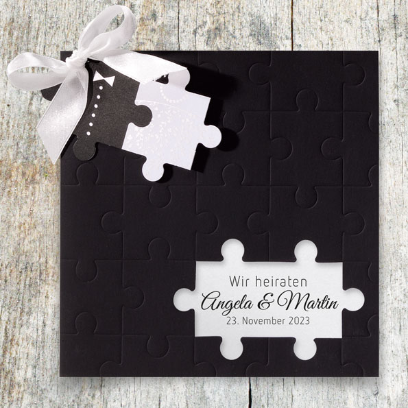 Puzzle-Hochzeitseinladungen – vergrößerte Abbildung Schwarze Einladungskarte zur Hochzeit, an die mit einer weißen Schleife zwei Puzzleteile angeknüpft sind.